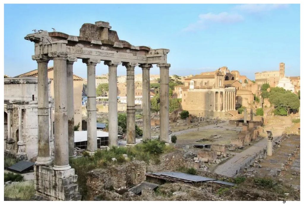 A full-slide color photograph of the Roman Forum in present-day Rome, viewed from an elevated vantage point. In the foreground, broken marble columns, temple fragments, stone foundations, and ancient roads spread across the archaeological site. A broad path leads the eye through the ruins toward the distance, where churches, Renaissance and later buildings, clusters of trees, and low hills reveal the living city beyond. Warm, golden light bathes the pale stone, emphasizing Rome's layers of history. From the Antiquity and the Renaissance Bridge lecture by Ernesto.